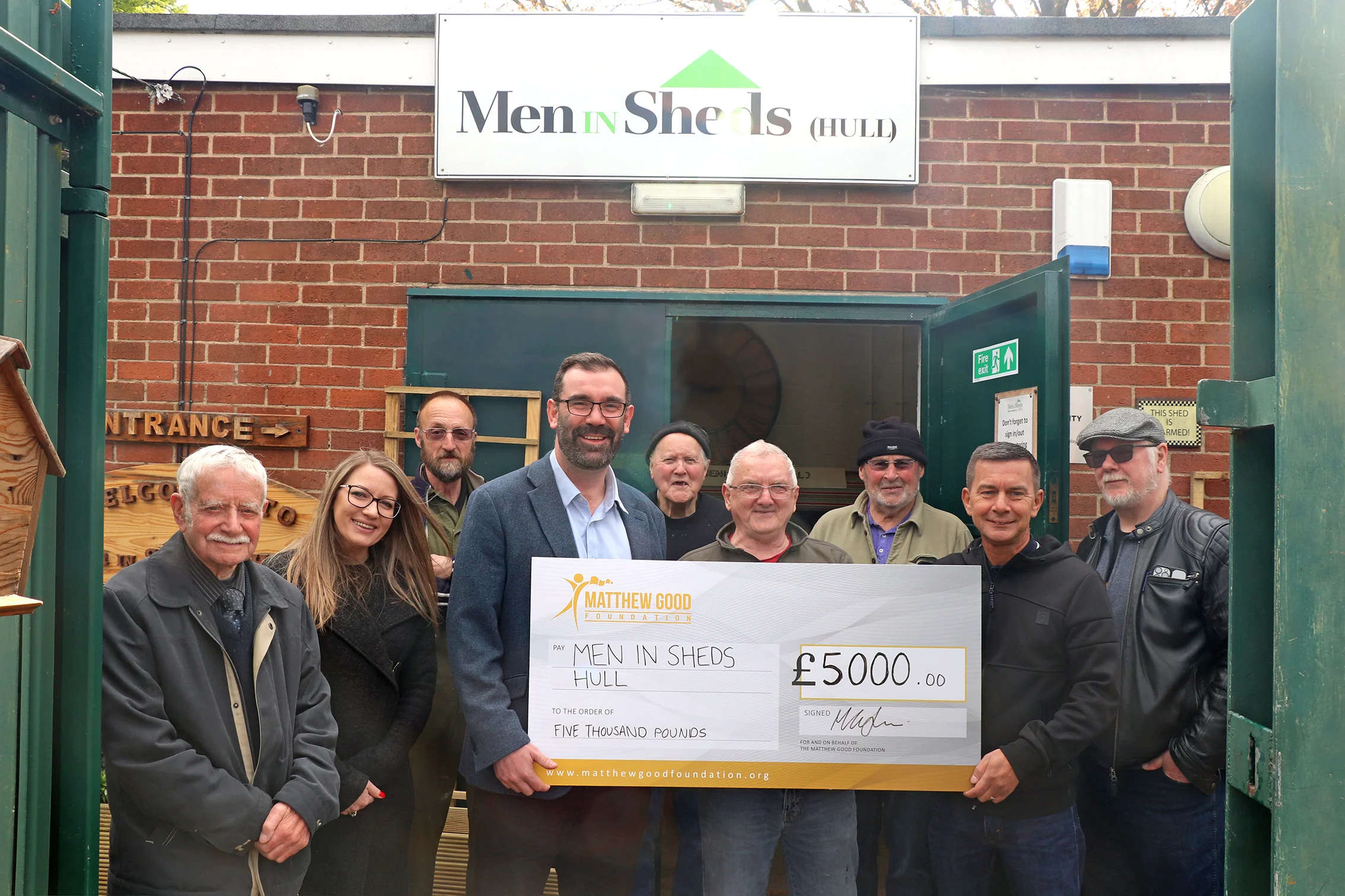Adam Walsh, CEO of John Good group and Michelle Taft, Executive Director of The Matthew Good Foundation hand over a cheque for £5,000 to Nick Todd - manager of Men in Sheds Hull. a group of service users are standing behind them